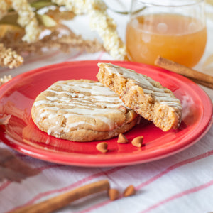 A close-up of an Apple Cider Cookie and a half cookie resting on a plate, surrounded by cinnamon chips. A glass of apple cider and cinnamon sticks sit alongside autumn leaves and fall foliage in the background, creating a cozy autumn scene.