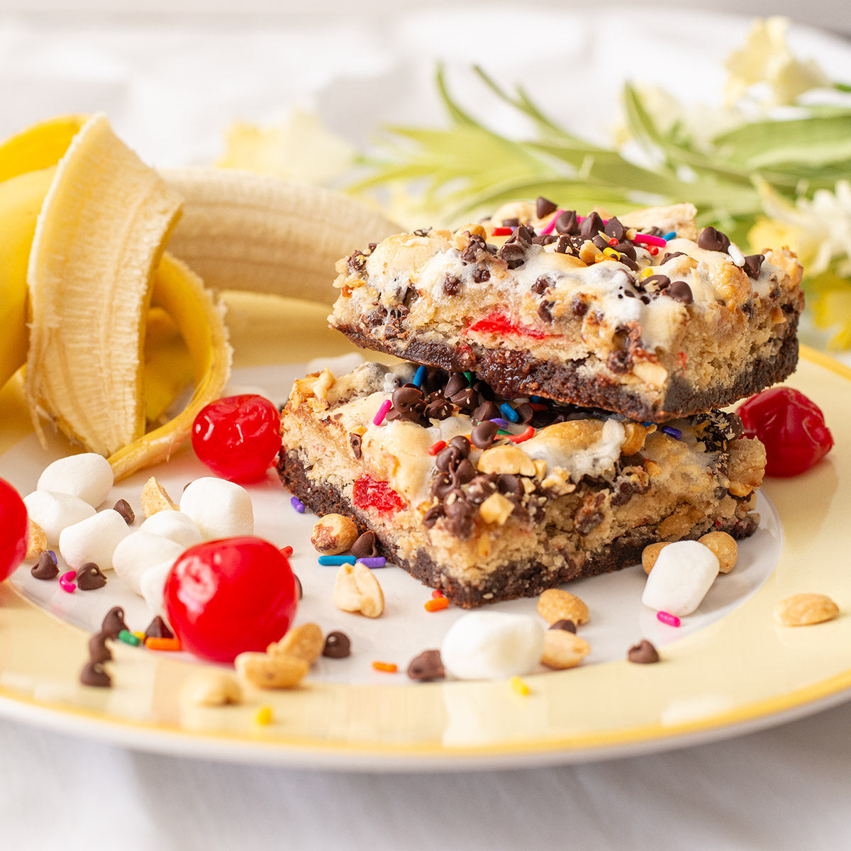 A tantalizing display of Banana Split Brownies arranged on a plate, adorned with a colorful assortment of marshmallows, chocolate chips, sprinkles, maraschino cherries, and peanuts. In the background, a fresh banana adds to the vibrant and delicious composition, inviting you to enjoy a taste of this delightful treat packed with summer-inspired flavors.