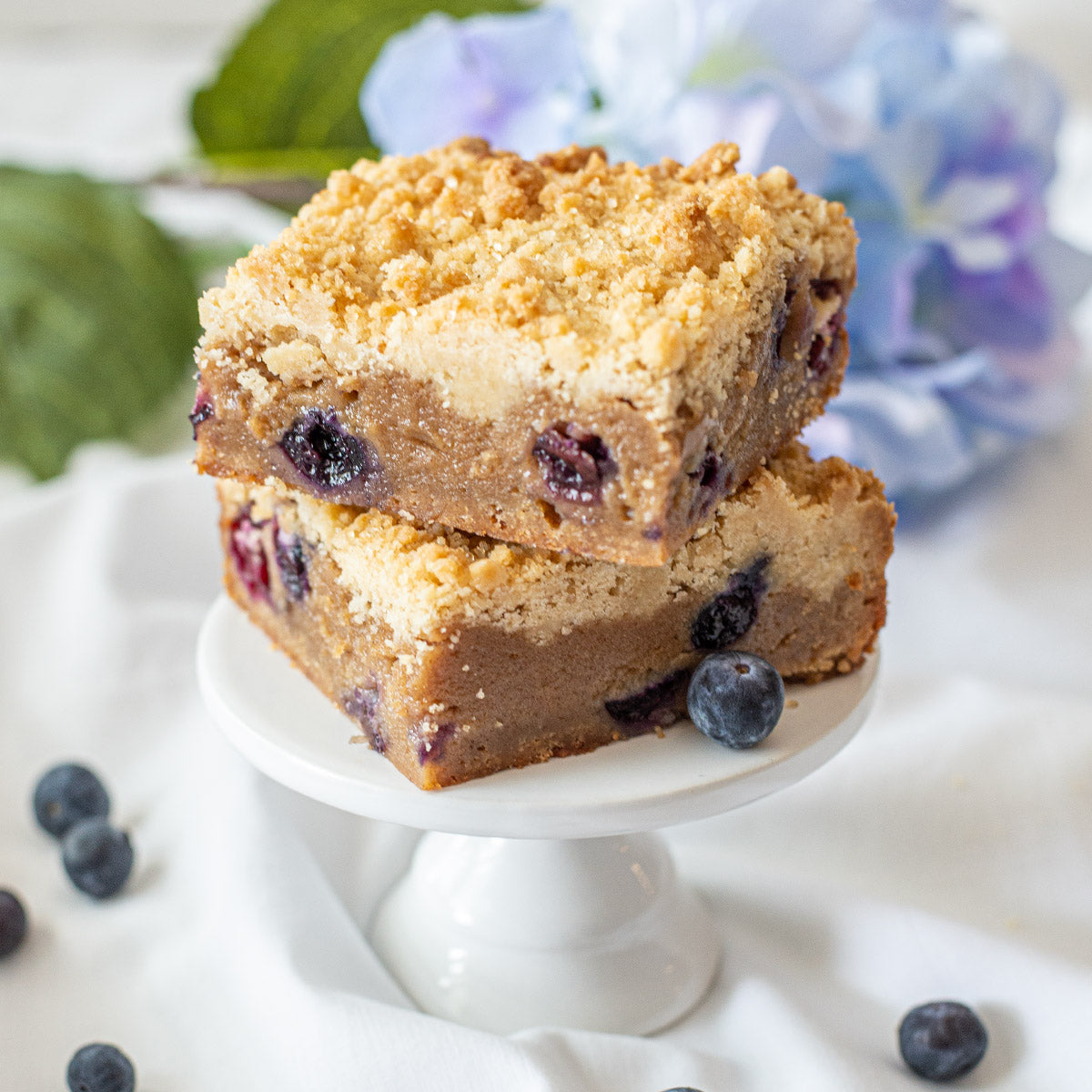 Two Blueberry Crumb Blondie bars stacked on a stand, surrounded by fresh blueberries, showcasing their delicious fruity flavor.
