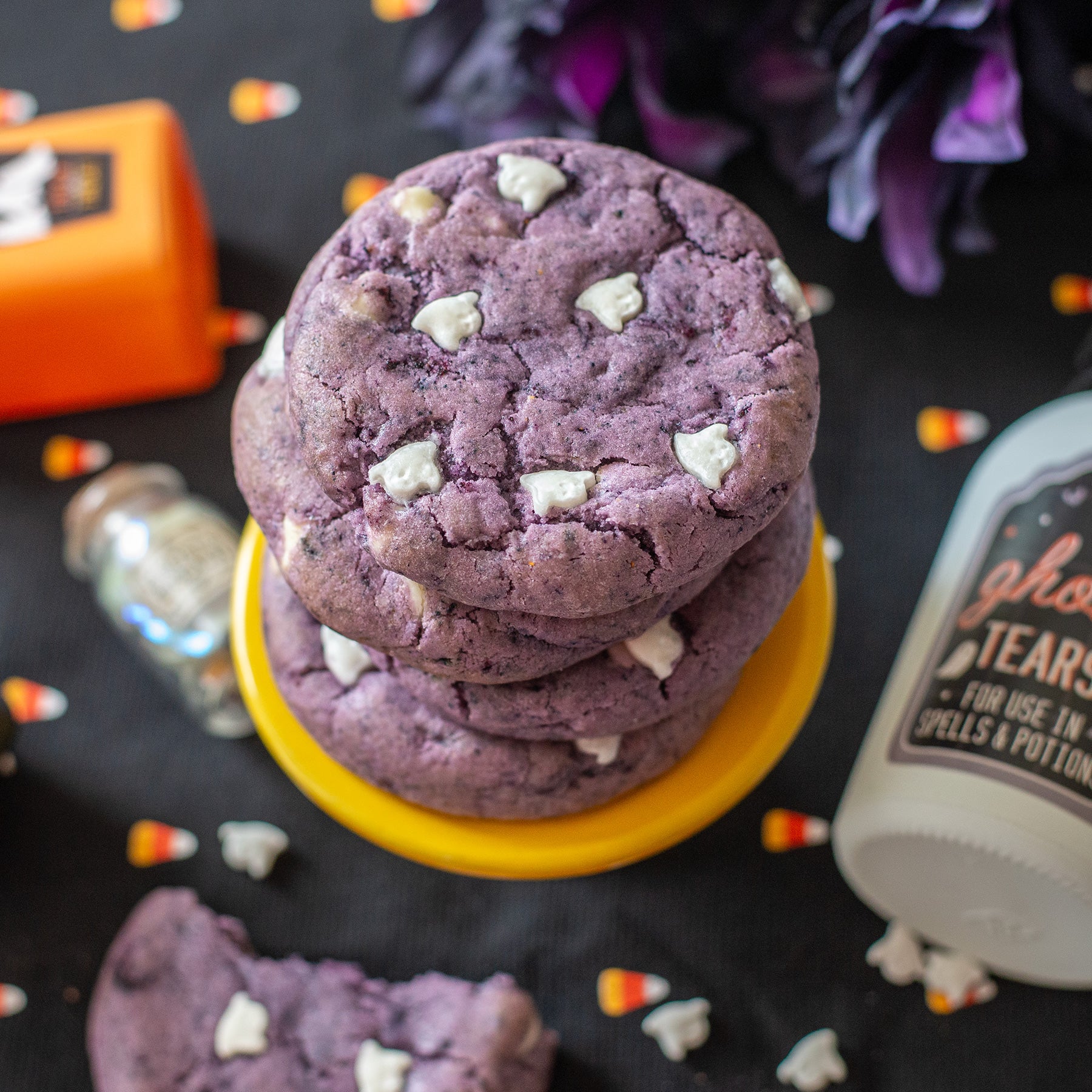 Stack of Boo-Berry cookies with ghost  sprinkles on a black surface with Halloween-themed decorations.