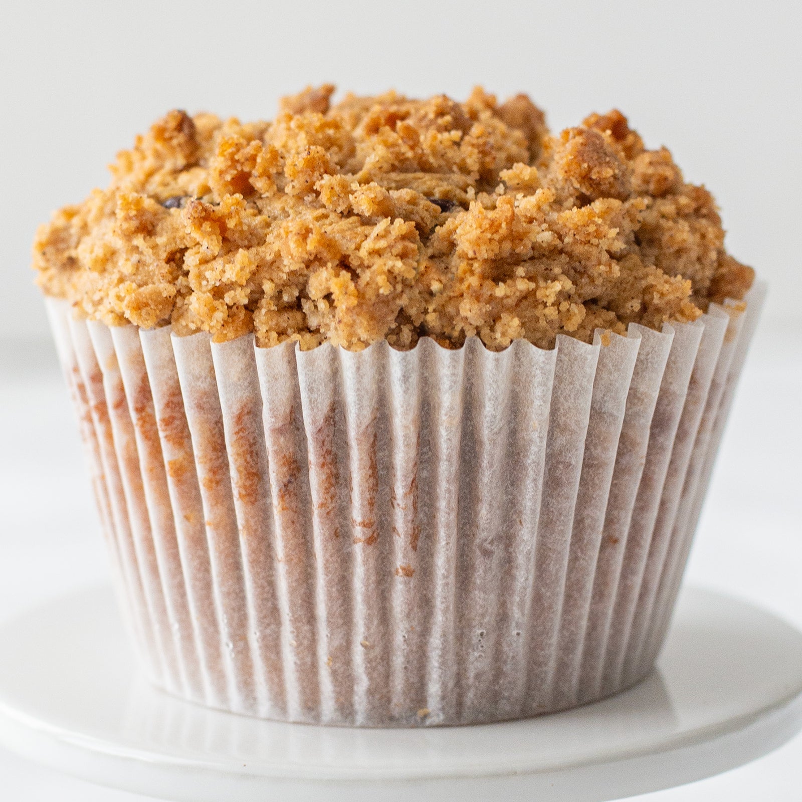 Single cappuccino muffin on a pedestal stand, topped with a golden-brown cinnamon streusel and visible chocolate chips, set against a softly lit background.