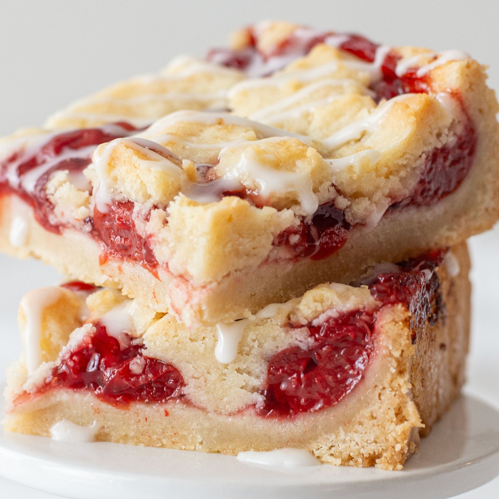 Overhead shot of a cherry pie bar on a plate, showing layers of golden shortbread crust, vibrant cherry filling, crumb topping, and a light almond glaze drizzle.