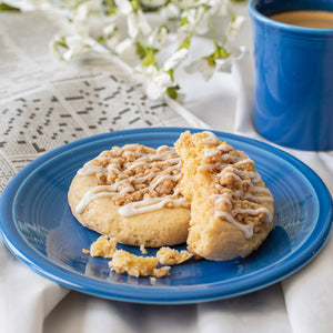 Broken Crumb Cake Cookie on a plate next to a cup of coffee and a newspaper crossword puzzle in the background. The cookie shows its crumbly streusel topping and soft interior.