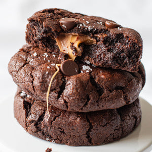 An up-close photo of a freshly baked Salted Caramel Cookie, highlighting the gooey caramel center encased within the chocolate cookie dough, and the elegant touch of fleur de sel on top.