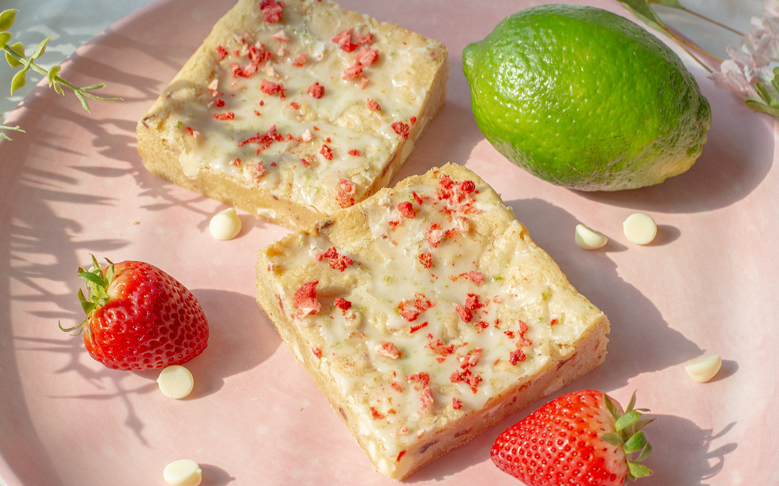 Two Strawberry Lime Blondies on a plate surrounded by scattered chocolate chips, strawberries and a lime.