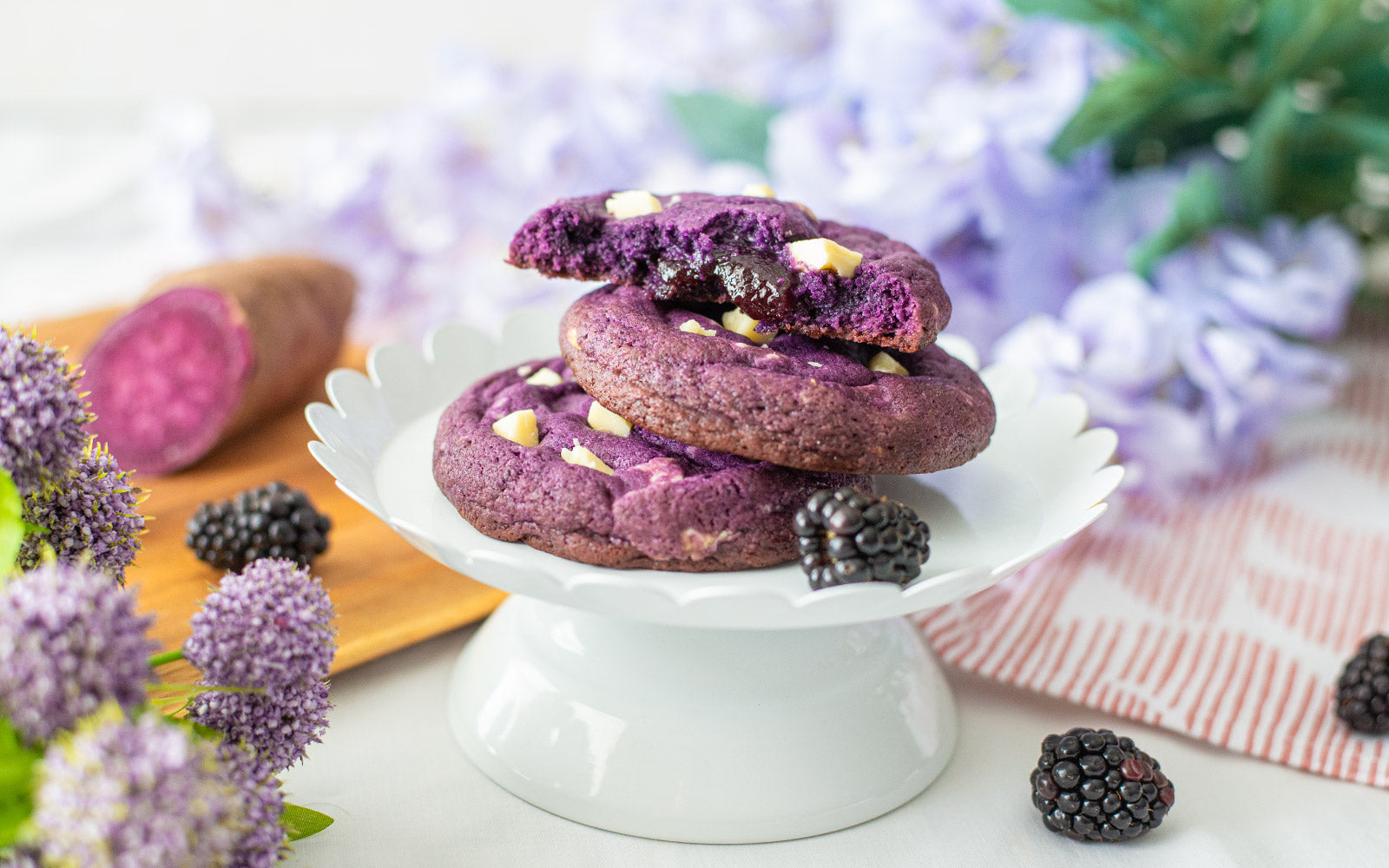 A stack of Ube Blackberry Cookies on a plate surrounded by purple yam, blackberries and flowers.