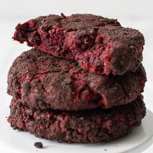 Three stacked Velvet Vortex Cookies, with the top cookie broken in half to reveal the rich red velvet interior, mini chocolate chips, and the contrasting dark Oreo crust around the edges.